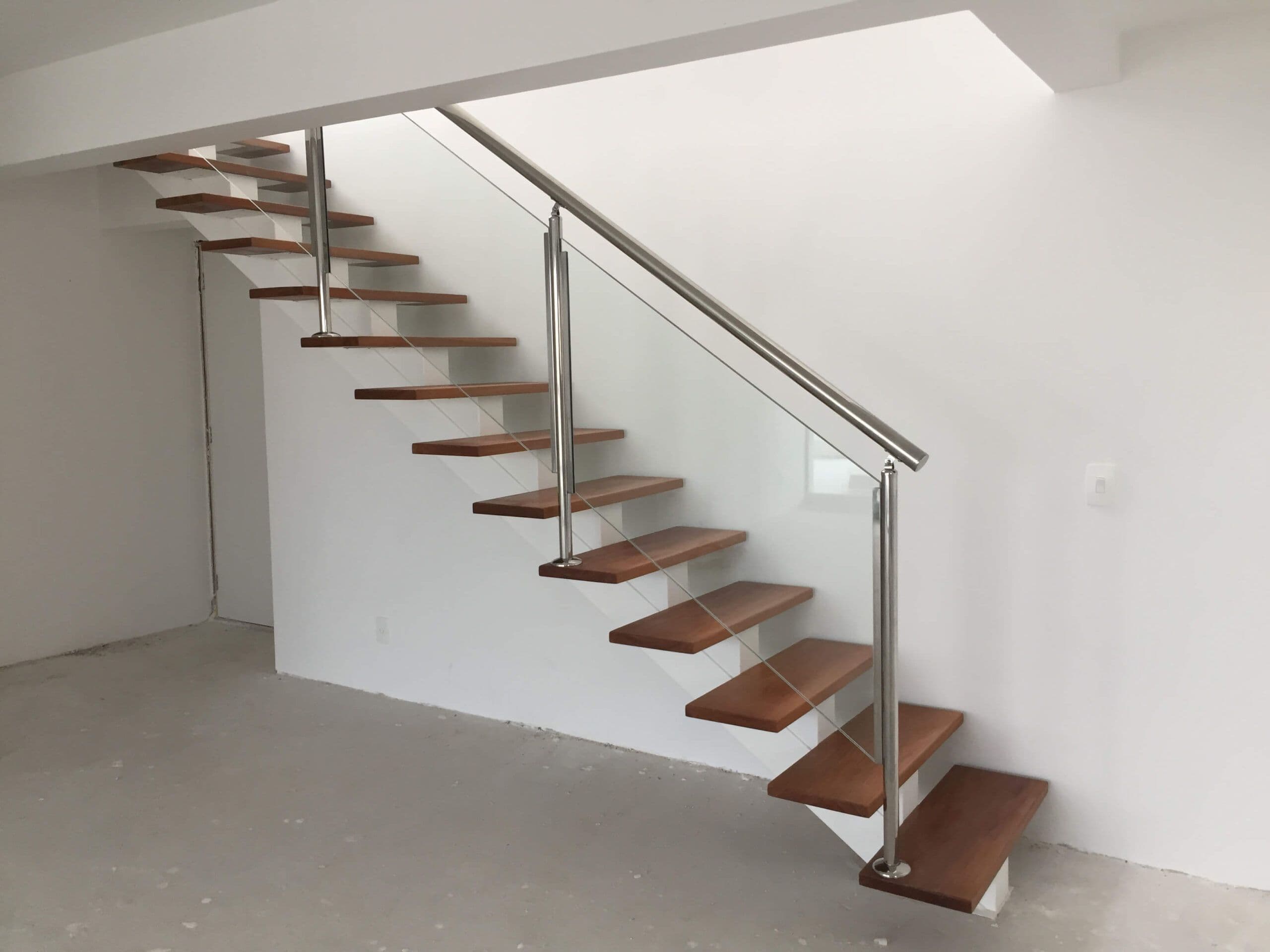 Escada reta com degraus de madeira, guarda-corpo de vidro e corrimão de inox em ambiente ainda em obra